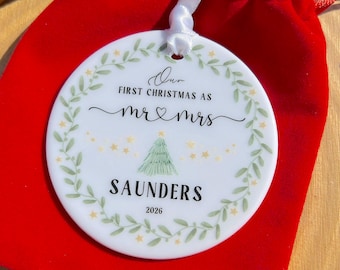 May include: A round white ceramic ornament featuring a green leaf wreath border, a small green Christmas tree, and yellow stars. The text reads: Our FIRST CHRISTMAS AS mr & mrs SAUNDERS 2026. A white ribbon is attached to the top for hanging.