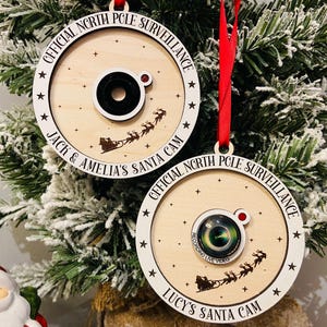 May include: Two round wooden ornaments with white text and stars. The ornaments are decorated with a black camera lens and a red dot. The text on the ornaments reads "Official North Pole Surveillance Jack & Amelia's Santa Cam" and "Official North Pole Surveillance Lucy's Santa Cam".