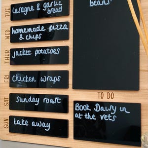 May include: A wooden weekly meal planner with black chalkboard labels. The labels are written in white chalk with a list of meals for each day of the week. A separate chalkboard label is used for a to-do list with the text "Book Dally in at the vets".