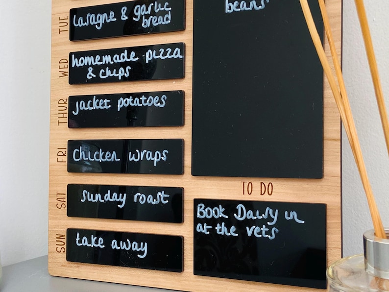 May include: A wooden weekly meal planner with black chalkboard labels. The labels are written in white chalk with a list of meals for each day of the week. A separate chalkboard label is used for a to-do list with the text "Book Dally in at the vets".