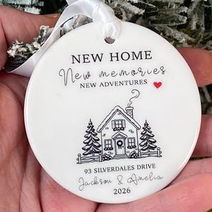 May include: A round white ceramic ornament with a white ribbon, featuring black text that reads NEW HOME, New memories, NEW ADVENTURES, 93 SILVERDALES DRIVE, Jackson & Amelia, 2026, a small red heart, and a black line drawing of a house.