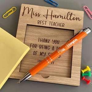 May include: A wooden picture frame engraved with "Miss Hamilton BEST TEACHER" and an orange pen with "Miss Hampton" printed on it. There are also yellow sticky notes, paperclips and push pins in the image.