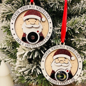 May include: Two round wooden ornaments with red ribbon hangers. Each ornament features a white Santa Claus with a red hat and a glitter beard holding a camera. The text on the ornaments reads "Official North Pole Surveillance" and "Jack & Amelia's Santa Cam" and "Oliver's Santa Cam".