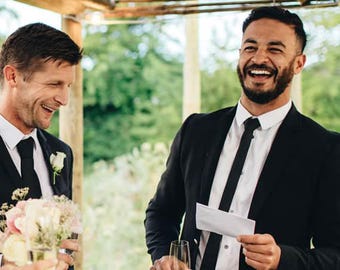 Op de afbeelding: Een bruiloftscène in de buitenlucht met drie lachende mensen. Een persoon in een zwart pak en stropdas lacht en houdt een papier vast. Een persoon in een witte trouwjurk en een persoon in een zwart pak glimlachen ook. Een boeket bloemen is zichtbaar.