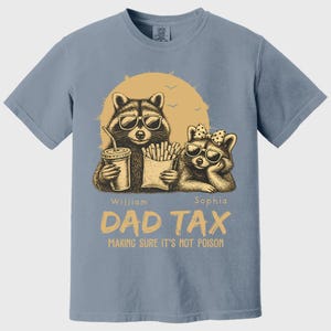 May include: Pale blue t-shirt with a graphic of two raccoons in sunglasses, holding a drink and fries, and the text "DAD TAX MAKING SURE IT'S NOT POISON". The shirt has a Comfort Colors tag.