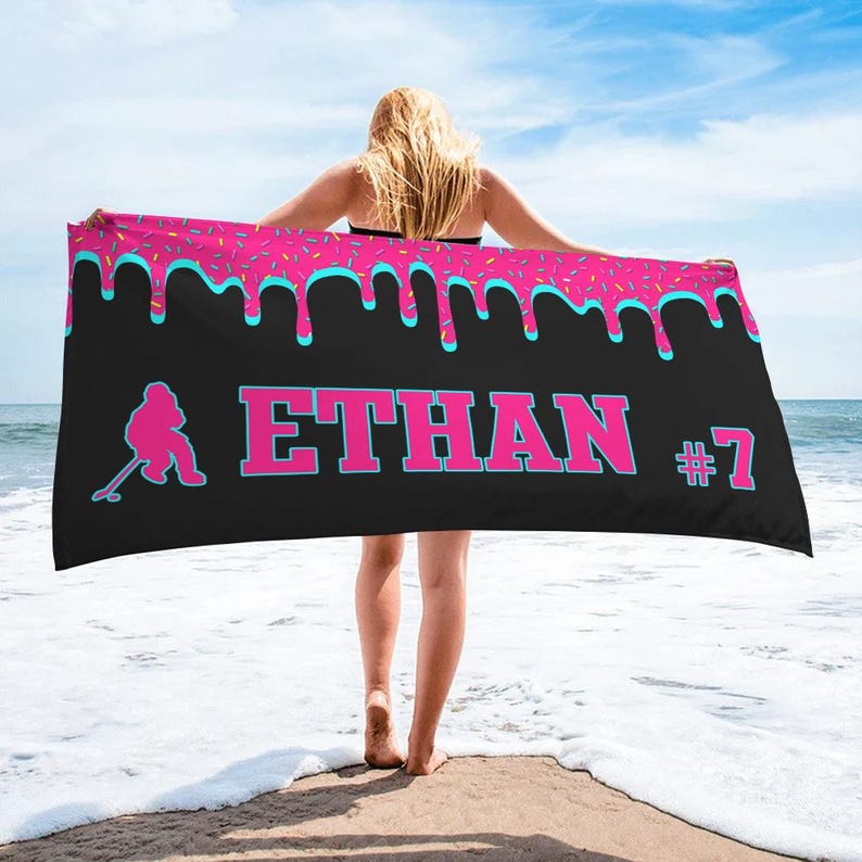 May include: A black beach towel with a pink and blue dripping icing design. The towel has the name "ETHAN #7" printed on it in pink letters. A silhouette of a hockey player is on the left side of the towel.
