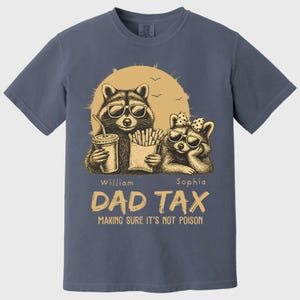 May include: Blue t-shirt with a graphic of two raccoons wearing sunglasses, set against a sun. One raccoon holds a drink, the other fries. Text reads "DAD TAX MAKING SURE IT'S NOT POISON."