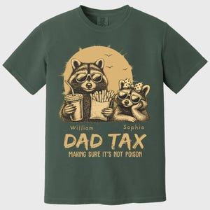May include: Dark green t-shirt with a graphic of two raccoons wearing sunglasses. The text reads "DAD TAX MAKING SURE IT'S NOT POISON". One raccoon holds a drink, the other holds fries. The names "William" and "Sophia" are below the raccoons.