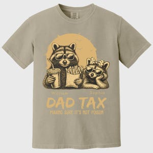 May include: Tan t-shirt with a graphic of two raccoons wearing sunglasses, holding a drink and fries. The text "DAD TAX" and "MAKING SURE IT'S NOT POISON" are printed below the raccoons. The names "William" and "Sophia" are also printed.