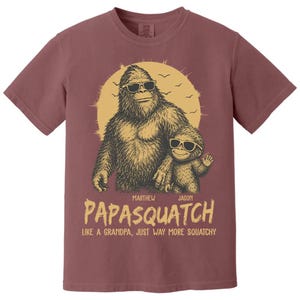 May include: A maroon t-shirt with a graphic of a Bigfoot-like creature and a smaller version, both wearing sunglasses. The text "PAPASQUATCH" is below the image, with the tagline "LIKE A GRANDPA, JUST WAY MORE SQUATCHY."