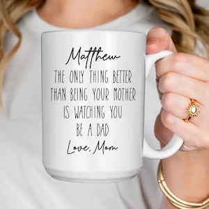 May include: White ceramic mug with the text "Matthew, The only thing better than being your mother is watching you be a dad, Love, Mom" in black script. The mug has a large handle and is held by a person.