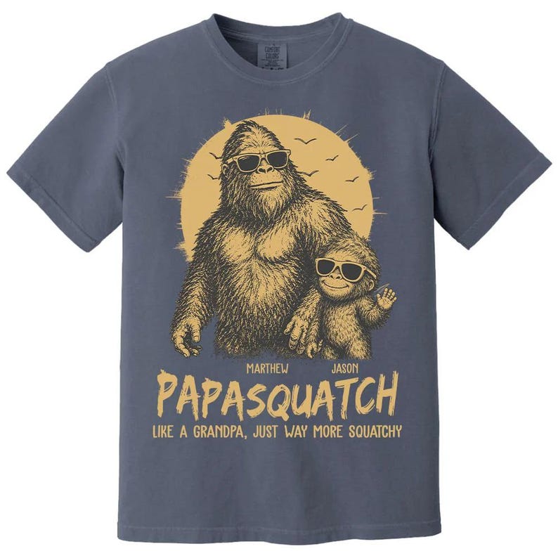 May include: Blue t-shirt with a Bigfoot and child graphic wearing sunglasses, set against a yellow sun. The text reads "PAPASQUATCH" and "LIKE A GRANDPA, JUST WAY MORE SQUATCHY". A fun, novelty design.