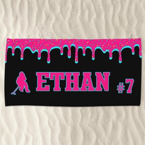 May include: Black beach towel with a pink and blue dripping icing design. The towel has a silhouette of a hockey player and the text "ETHAN #7".