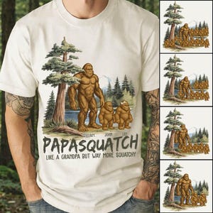 Può includere: T-shirt color crema con un'illustrazione della famiglia Bigfoot. Il design include una grande figura di Bigfoot con due più piccole, un lago, alberi e montagne. Il testo recita "PAPASQUATCH" e "LIKE A GRANDPA BUT WAY MORE SQUATCHY."