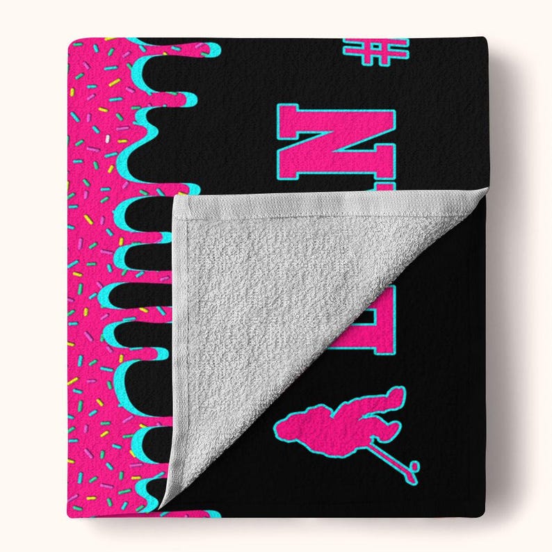 May include: Black bath towel with a pink and blue dripping icing design, the word "Ninja" in pink letters, and a pink stick figure holding a stick. The towel has a white terry cloth backing.