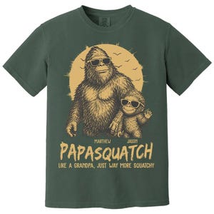 May include: Green t-shirt with a Bigfoot and child Bigfoot graphic wearing sunglasses, set against a yellow sun. The text reads "PAPASQUATCH" and "LIKE A GRANDPA, JUST WAY MORE SQUATCHY."
