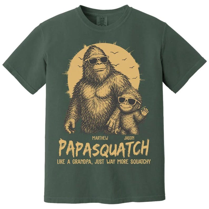 May include: Green t-shirt with a Bigfoot and child Bigfoot graphic wearing sunglasses, set against a yellow sun. The text reads "PAPASQUATCH" and "LIKE A GRANDPA, JUST WAY MORE SQUATCHY."