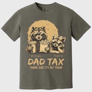 May include: A grey t-shirt with a graphic of two raccoons wearing sunglasses, holding a drink and fries, with the text "DAD TAX" and "MAKING SURE IT'S NOT POISON". The shirt has a vintage, distressed appearance.