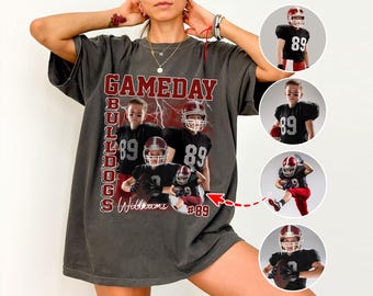 May include: Dark gray t-shirt featuring a football-themed design. The shirt displays the text "GAMEDAY BULLDOGS" in red and white, along with images of football players. The number "89" is prominently featured.