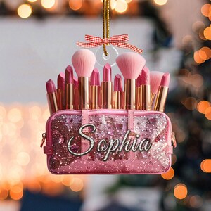May include: A pink cosmetic bag ornament with the name "Sophia" in cursive script. The bag is filled with pink lipsticks and makeup brushes. A red and white ribbon is attached for hanging.