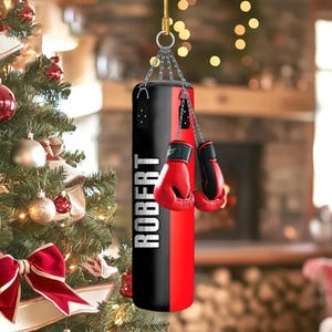 May include: A black and red boxing bag with the name "ROBERT" printed vertically in white. Red boxing gloves hang from the bag. The bag is suspended by a chain. The background includes a Christmas tree and a fireplace.