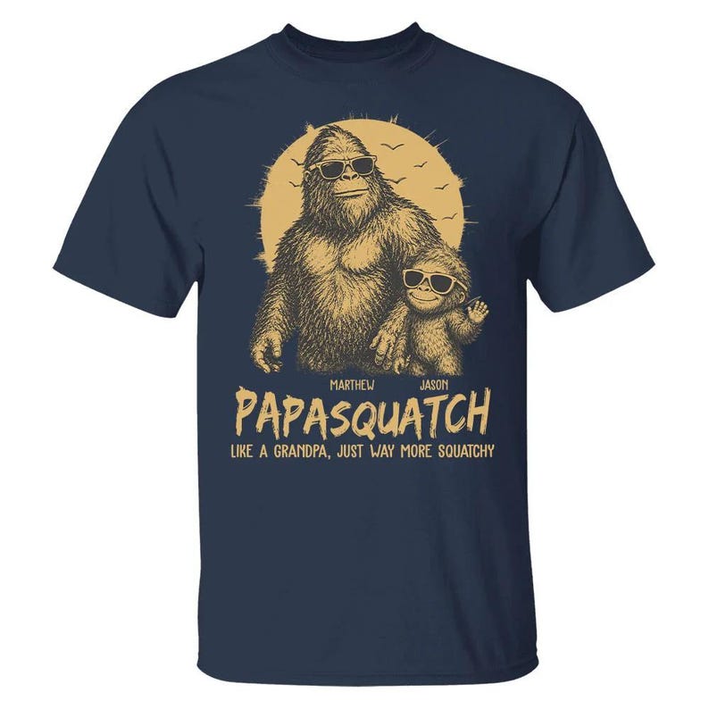 May include: Navy blue t-shirt with a Bigfoot and child graphic wearing sunglasses, set against a sun. The text says "Papasquatch" and "Like a grandpa, just way more squatchy". The design is in brown and tan.