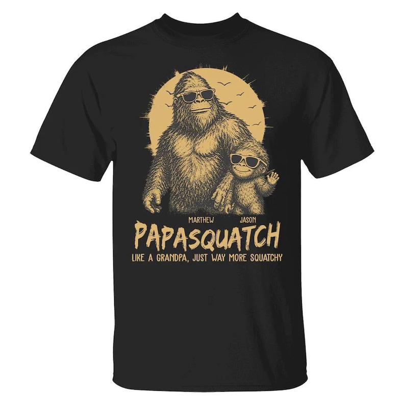 May include: Black t-shirt with a Bigfoot and child graphic wearing sunglasses, set against a yellow sun and birds. The text says "Papasquatch", with names "Marthew" and "Jason", and the slogan "Like a grandpa, just way more squatchy".