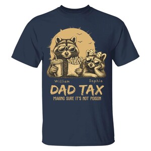 May include: Navy blue t-shirt with a graphic of two raccoons wearing sunglasses, holding a drink and fries. The text reads "DAD TAX" and "MAKING SURE IT'S NOT POISON". The design includes a sun and birds.