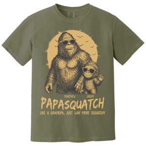 May include: Khaki green t-shirt with a Bigfoot and child graphic wearing sunglasses. The text says "PAPASQUATCH" with names "Marthew" and "Jason" above and "LIKE A GRANDPA, JUST WAY MORE SQUATCHY" below the image. A fun, novelty tee.