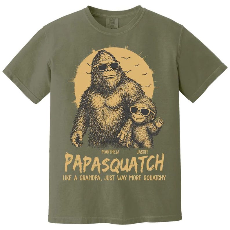 May include: Khaki green t-shirt with a Bigfoot and child graphic wearing sunglasses. The text says "PAPASQUATCH" with names "Marthew" and "Jason" above and "LIKE A GRANDPA, JUST WAY MORE SQUATCHY" below the image. A fun, novelty tee.