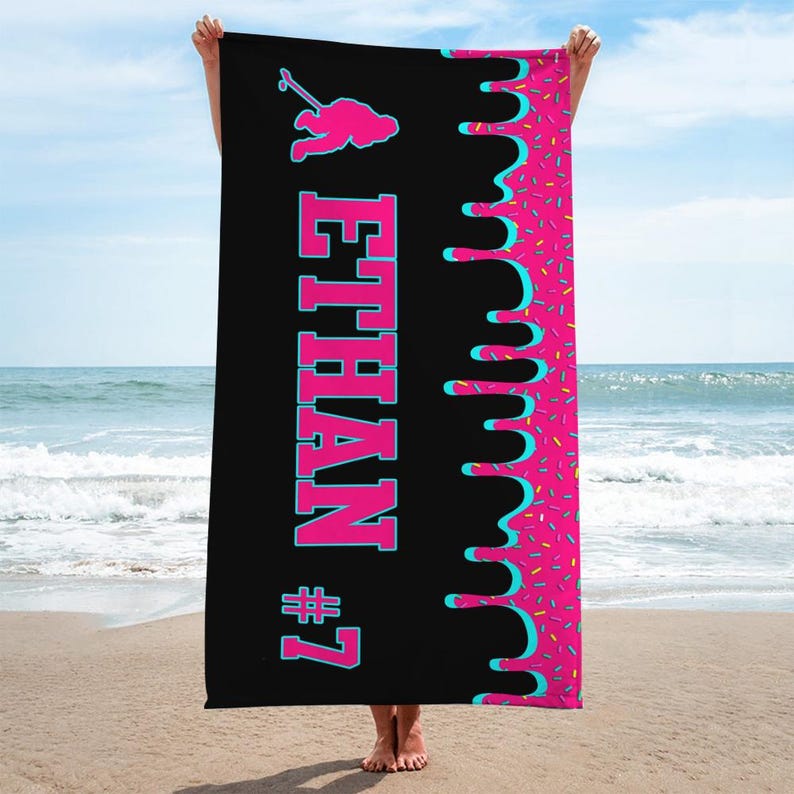 May include: Black beach towel with a pink and blue dripping glaze design. The towel has the name "ETHAN #7" in pink and blue lettering.