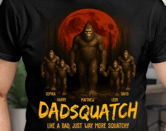 May include: Black t-shirt featuring a Bigfoot family illustration under a red moon. The design includes the text "Dadsquatch" in yellow, with the tagline "Like a Dad, Just Way More Squatchy." Names are printed below the Bigfoot figures.