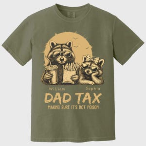 May include: Olive green t-shirt with a graphic of two raccoons wearing sunglasses, set against a sun and birds. One raccoon holds a drink, the other fries. Text reads "DAD TAX MAKING SURE IT'S NOT POISON."