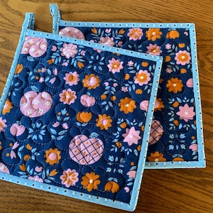 May include: Two quilted pot holders with a navy blue background and a floral pattern in pink and orange. The pot holders have a light blue border with white polka dots and a loop for hanging.