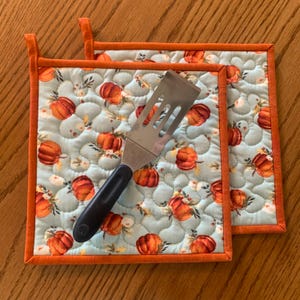 May include: Two square quilted pot holders with a light blue background and a pattern of orange pumpkins and floral accents. The pot holders have orange trim and hanging loops. A stainless steel spatula with a black handle rests on top.
