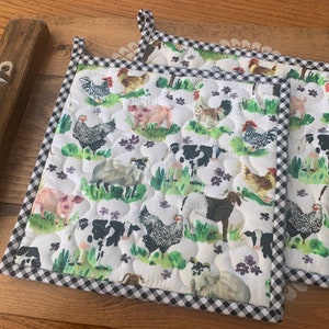 May include: Two quilted pot holders with a farm animal print featuring cows, chickens, pigs, and sheep. The pot holders have a black and white checkered border and a loop for hanging.