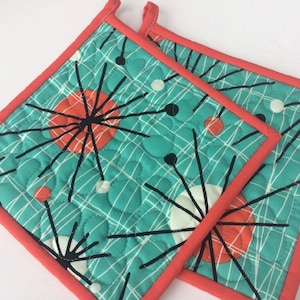 May include: Two square quilted pot holders with a teal background featuring a mid-century modern design. The design includes orange circles, black lines, and white dots. The edges are trimmed with a coral-colored binding.