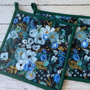 May include: Two square pot holders with a floral pattern in shades of blue, white, and gold on a black background. The edges are trimmed with a dark green binding, and each has a loop for hanging.