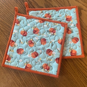 May include: Two square quilted pot holders featuring a light blue background with a repeating pattern of orange pumpkins and small white flowers. Each pot holder has a solid orange fabric border and a small fabric loop at the top corner.