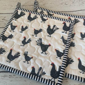 May include: Two quilted pot holders with a repeating pattern of black chickens with red combs on a white background. The pot holders have a black and white striped border and hanging loop.