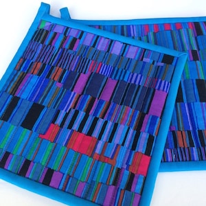 May include: Two square quilted potholders with a vibrant abstract design. The potholders feature a patchwork of vertical stripes in shades of blue, purple, black, and red, framed by a bright blue border. Each potholder has a small loop for hanging.