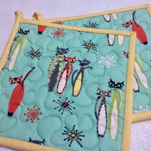 May include: Two square quilted pot holders with a light teal background. The fabric features a repeating pattern of stylized cats in red, black, and yellow, along with starburst and atomic shapes. The edges are trimmed with yellow binding.