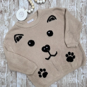 Puede incluir: Un suéter de punto beige con una cara de perro bordada en negro y huellas de patas. El suéter tiene cuello redondo y mangas largas.