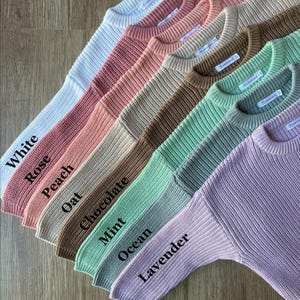 May include: A collection of ten sweaters in various colours, including white, rose, peach, oat, chocolate, mint, ocean, and lavender. The sweaters are arranged in a row, with the white sweater at the top and the lavender sweater at the bottom.
