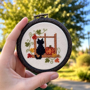 May include: A circular embroidery hoop with a dark wood-grain frame containing a cross-stitch design of a black cat sitting on a windowsill next to an orange pumpkin, surrounded by green and red autumn leaves on a cream-colored fabric background.