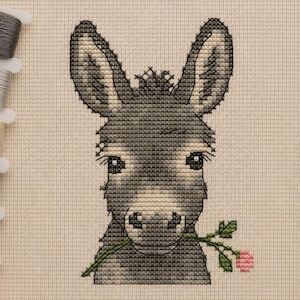 May include: A cross-stitch embroidery pattern on beige Aida fabric featuring a grey donkey with long ears holding a green stem with a small pink flower in its mouth, accompanied by a vertical row of six assorted embroidery thread bobbins.