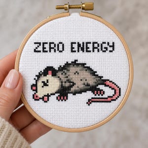 May include: A circular wooden embroidery hoop containing white Aida fabric with a cross-stitch design of a gray and white opossum. The text ZERO ENERGY is stitched in black block letters above the opossum, which has pink ears, feet, and tail.