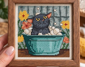 Könnte beinhalten: Ein gerahmtes Kreuzstichbild, das eine schwarze Katze in einer türkisfarbenen Waschschüssel voller weißer Seifenblasen zeigt. Der Hintergrund besteht aus einem Gittermuster mit gelben und orangefarbenen Blumen in einem quadratischen Holzrahmen.