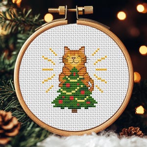May include: A cross-stitch embroidery hoop showing a smiling ginger cat embracing a Christmas tree. The tree is green with red baubles. The cat has yellow rays around it. The hoop is set against a blurred background of Christmas greenery and lights.