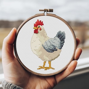 May include: A cross-stitch embroidery of a chicken in a wooden hoop. The chicken has a red comb and wattle, a yellow beak, and blue and white feathers. The hoop is held in a person's hand.